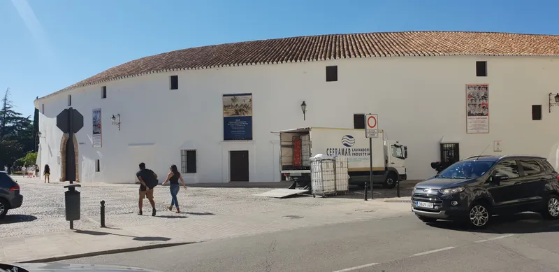 Plaza con un edificio blanco de tejado de teja, gente paseando y un coche aparcado