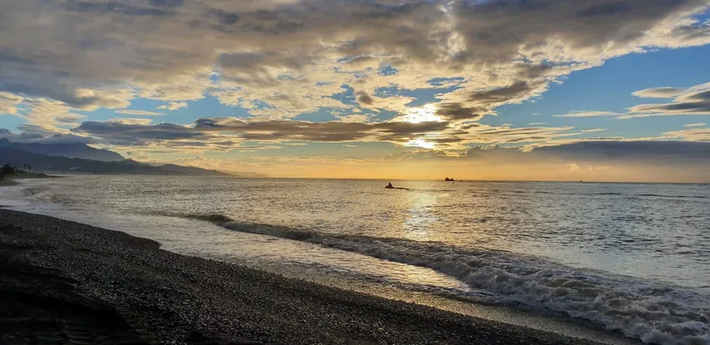 Olas llegando a una playa oscura bajo un cielo de nubes con el sol entre ellas