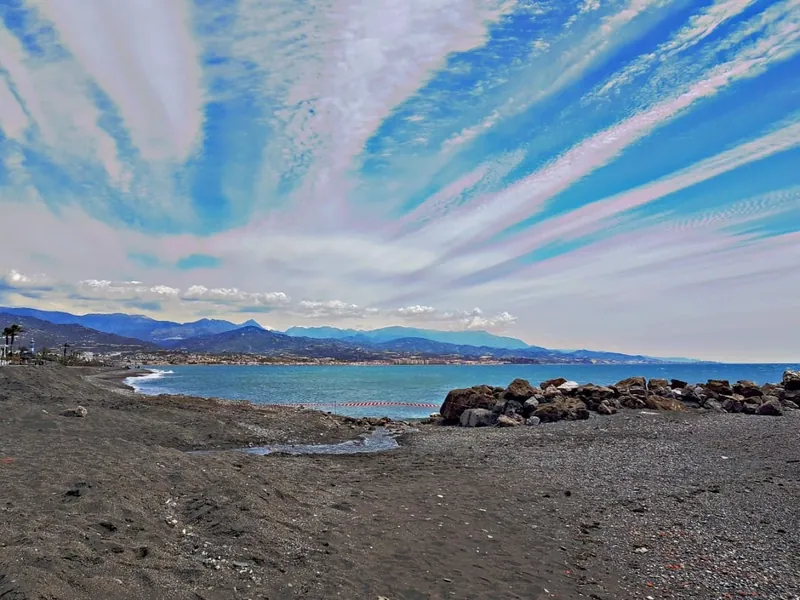 Cielo azul surcado por nubes alargadas sobre una playa con rocas y montañas
