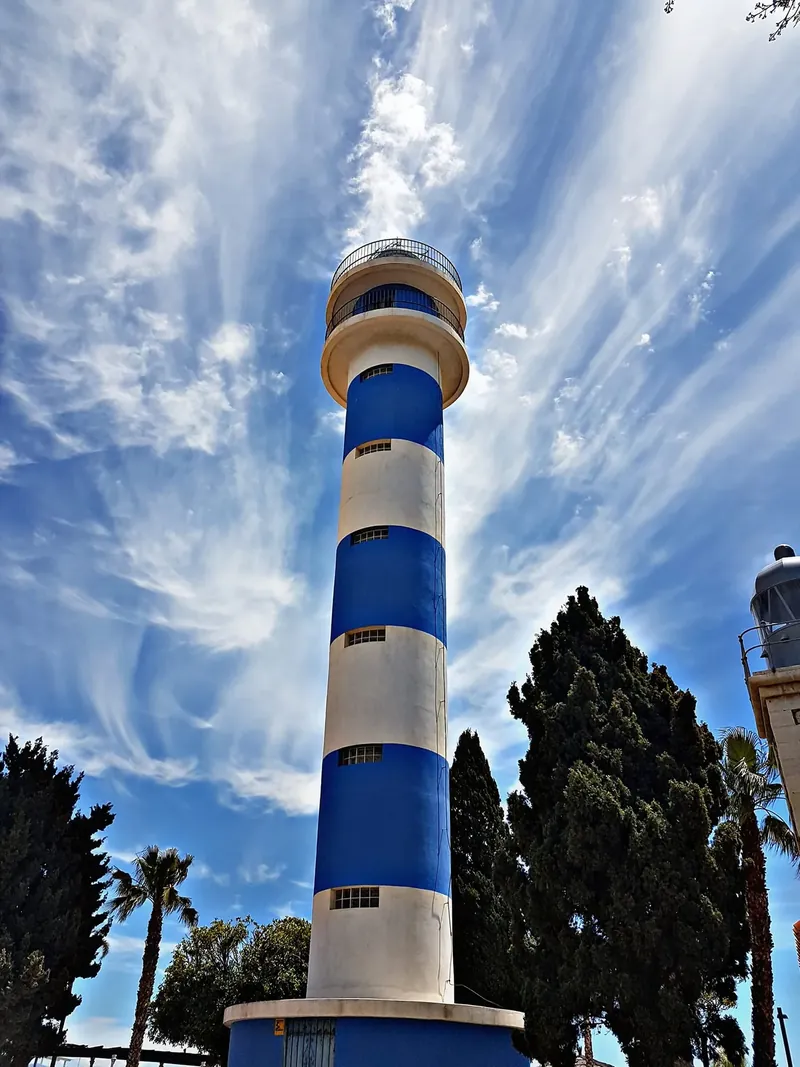 Faro de franjas azules y blancas con palmeras y un cielo de nubes finas
